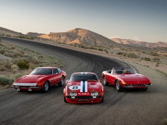 Three iconic Ferrari Daytona models parked on a winding desert road, set against a backdrop of arid mountains under a clear sky. From left to right: a red 1972 Ferrari 365 GTB/4 Daytona Berlinetta coupe, a red 1973 Ferrari 365 GTB/4 Daytona Competizione with NART racing livery and number 8, and a red 1973 Ferrari 365 GTS/4 Daytona Spider convertible with a tan interior.