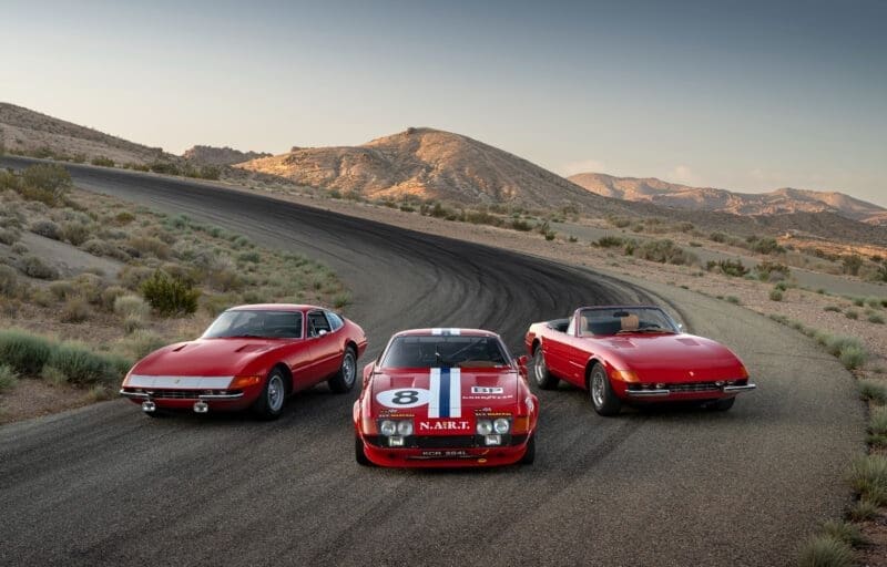 Three iconic Ferrari Daytona models parked on a winding desert road, set against a backdrop of arid mountains under a clear sky. From left to right: a red 1972 Ferrari 365 GTB/4 Daytona Berlinetta coupe, a red 1973 Ferrari 365 GTB/4 Daytona Competizione with NART racing livery and number 8, and a red 1973 Ferrari 365 GTS/4 Daytona Spider convertible with a tan interior.